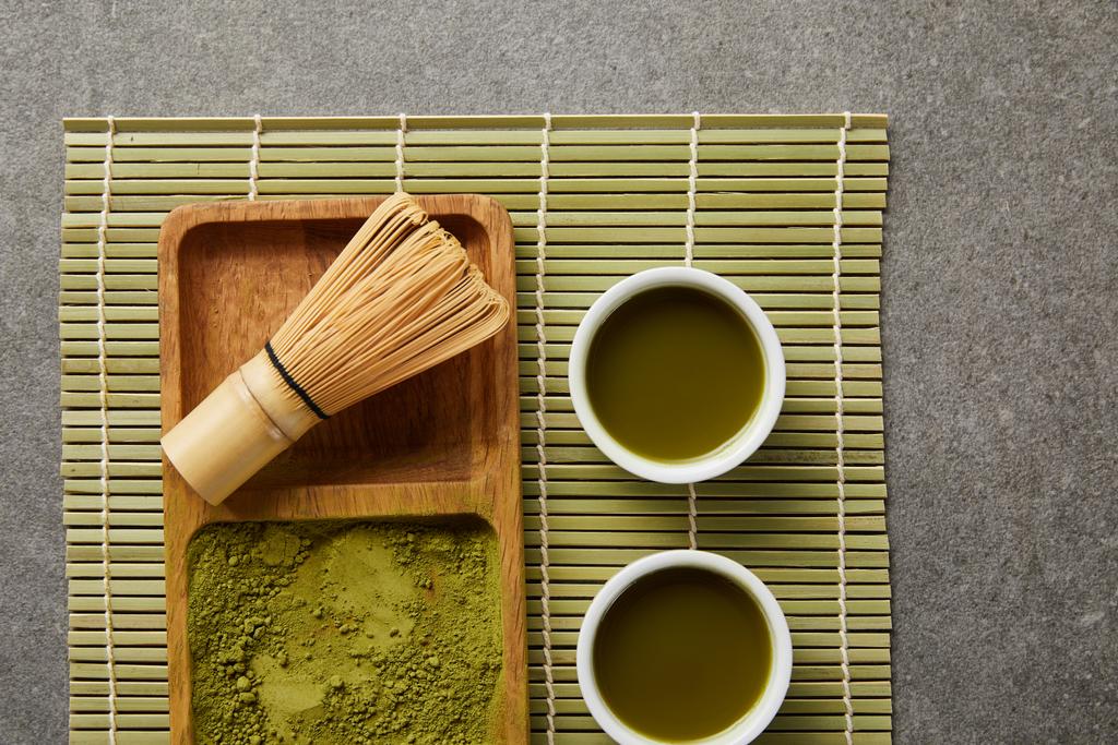 top view of green matcha powder with bamboo whisk on wooden board near white cups with green tea on table mat - Photo, Image
