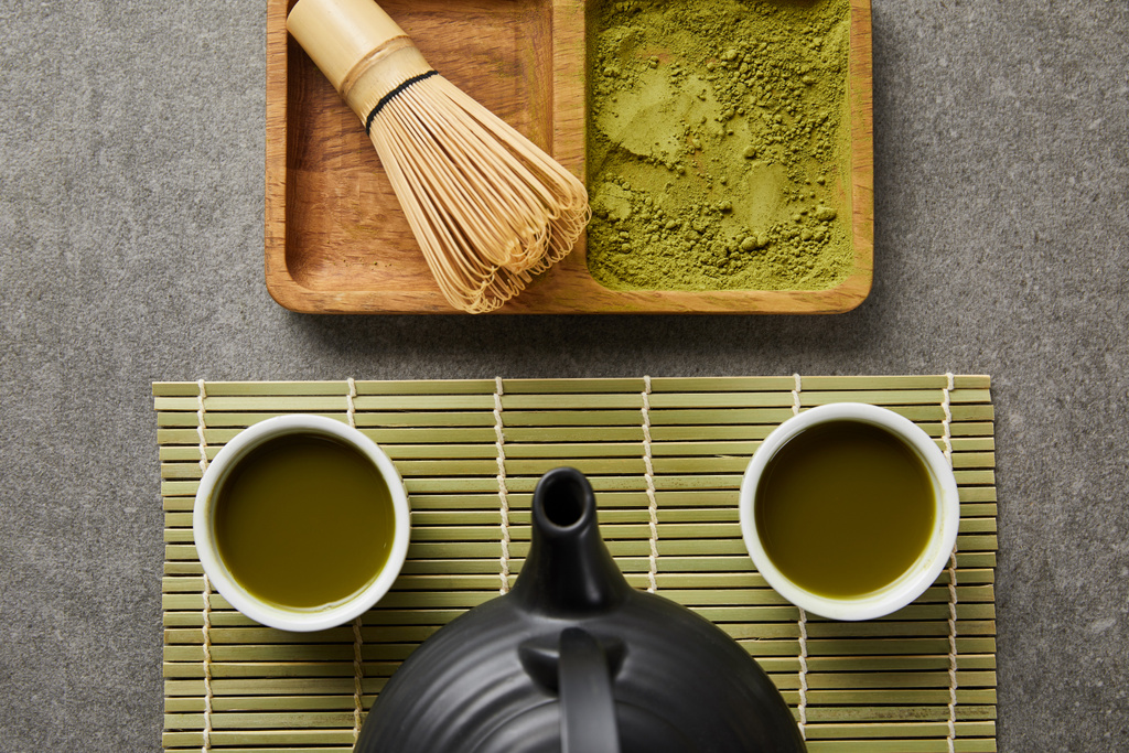 竹の泡立て器と抹茶パウダーの近くの緑のテーブルマットの上に紅茶と紅茶ポットと白いカップのトップビュー - 写真・画像