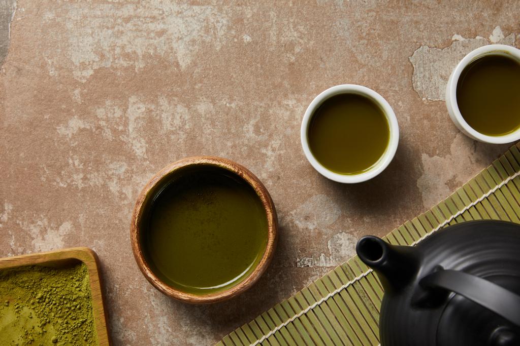 Top uitzicht van matcha poeder, Bowl en kopjes met groene thee op leeftijd oppervlak in de buurt van bamboe placemat en zwarte theepot - Foto, afbeelding