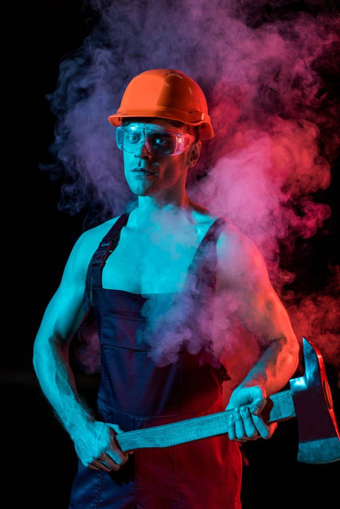 σοβαρός πυροσβέστης σε σκληρό καπέλο και προστατευτικά γυαλιά κρατώντας επίπεδη κεφάλι τσεκούρι σε καπνό σε μαύρο - Φωτογραφία, εικόνα