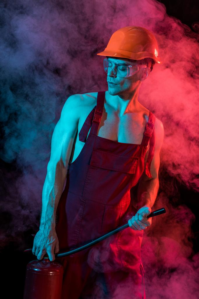 sexy hemdloser Feuerwehrmann im Overall hält Feuerlöscher in Rauch auf schwarz - Foto, Bild