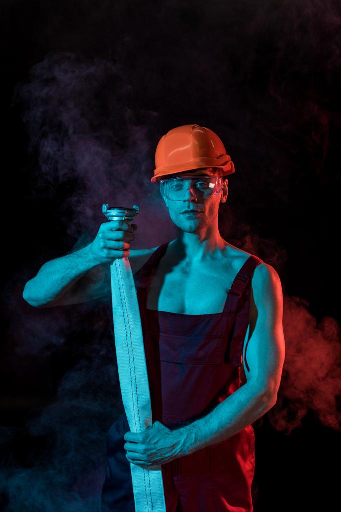 szexi tűzoltó a hardhat és védőszemüveg gazdaság tűzoltó tömlő füst fekete - Fotó, kép