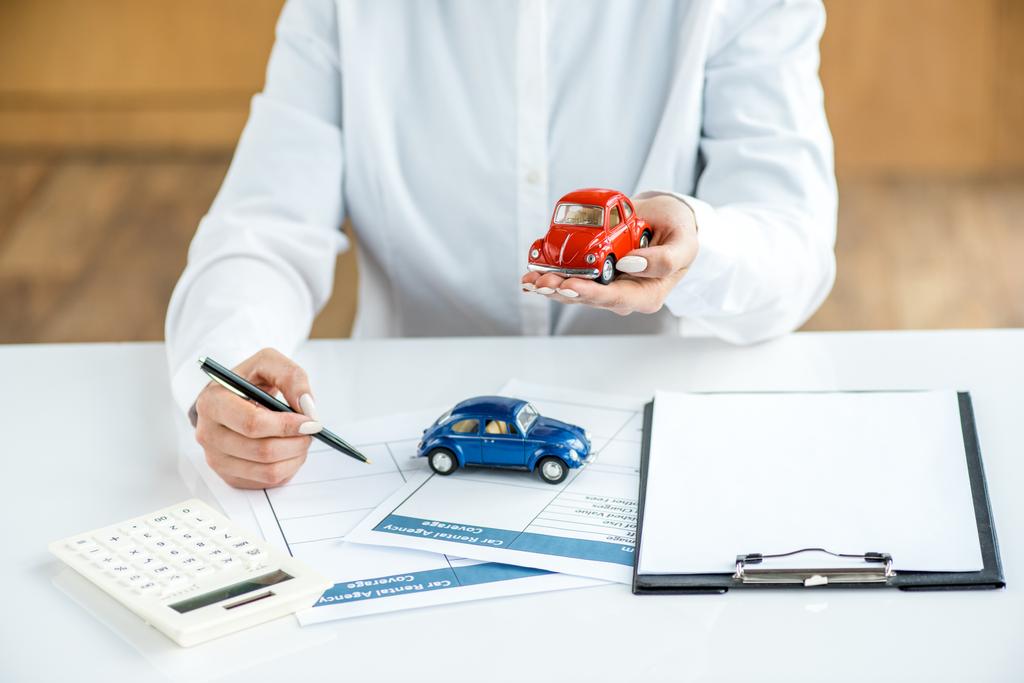 cropped view of woman in formal wear holding toy car and pen at table with documents, calculator and clipboard - Photo, Image
