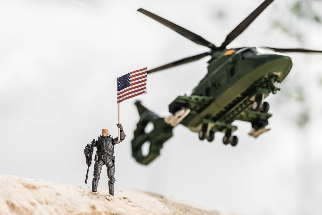 Spielzeugsoldat mit amerikanischer Flagge im Sand in der Nähe eines Hubschraubers - Foto, Bild