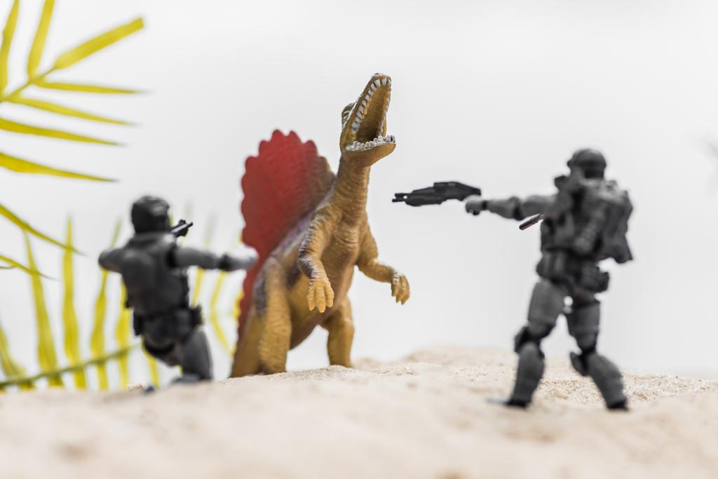 selektywne skupienie żołnierzy zabawek celowanie pistoletów na ryk maleńkiego dinozaura na piasku wzgórza - Zdjęcie, obraz