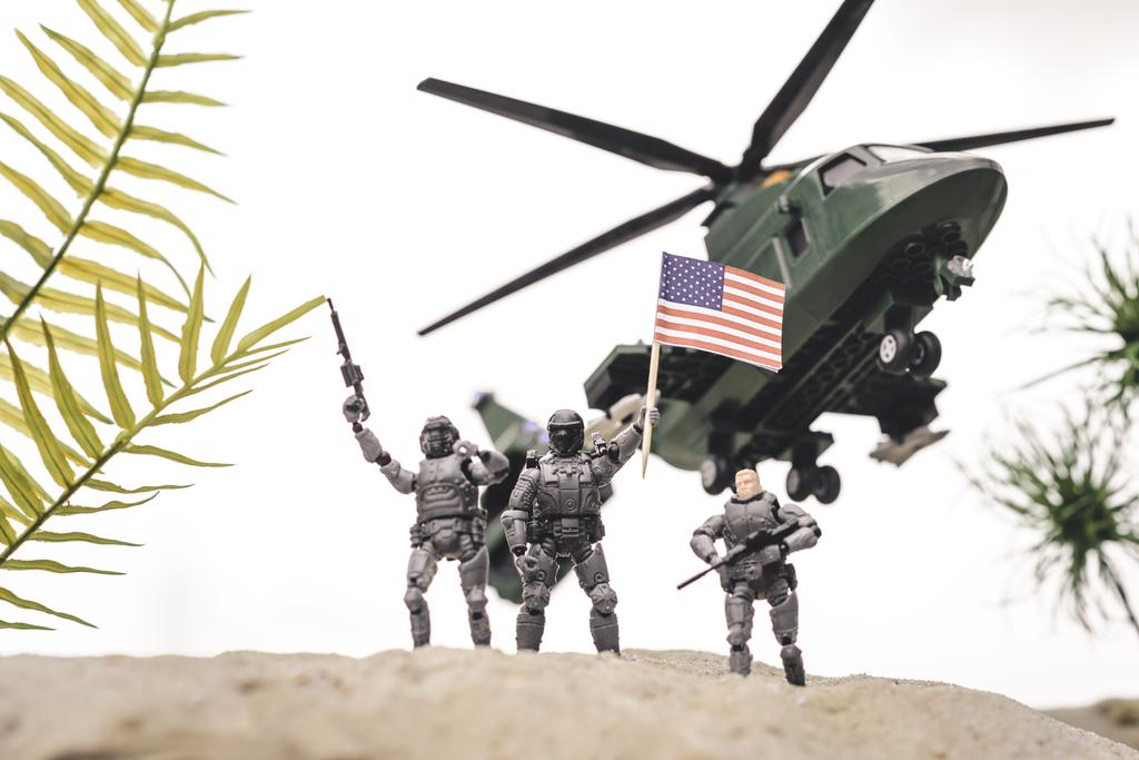 Spielzeugsoldaten aus Plastik auf Sanddüne mit Gewehren und amerikanischer Flagge in der Nähe von Hubschraubern am Himmel - Foto, Bild