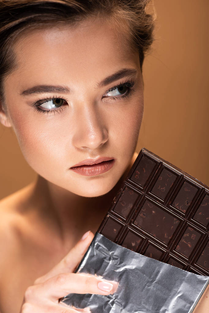 όμορφη νεαρή γυμνή γυναίκα κρατώντας μπάρα σοκολάτας σε ασημένιο φύλλο και κοιτάζοντας μακριά απομονωμένη σε μπεζ - Φωτογραφία, εικόνα