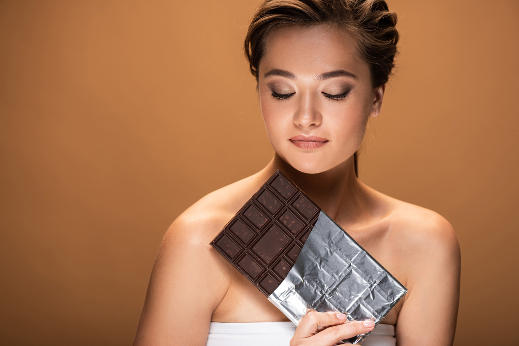 belle jeune femme aux yeux fermés tenant une barre de chocolat en feuille d'argent isolée sur beige
 - Photo, image