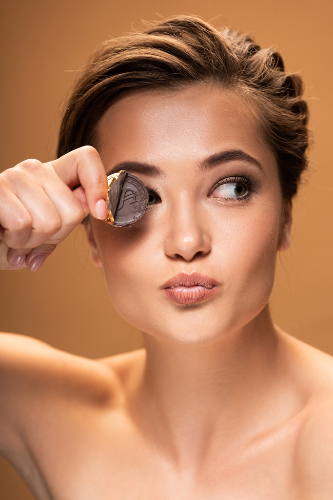 гола молода жінка тримає шоколадну монету в золотій фользі біля ока і дивиться ізольовано на бежевому
 - Фото, зображення