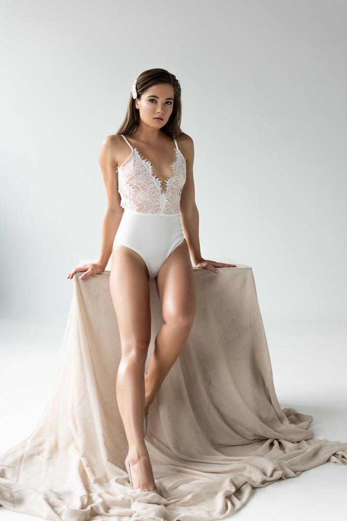 tender young woman in white lacy bodysuit sitting on beige cloth isolated on white - Photo, Image