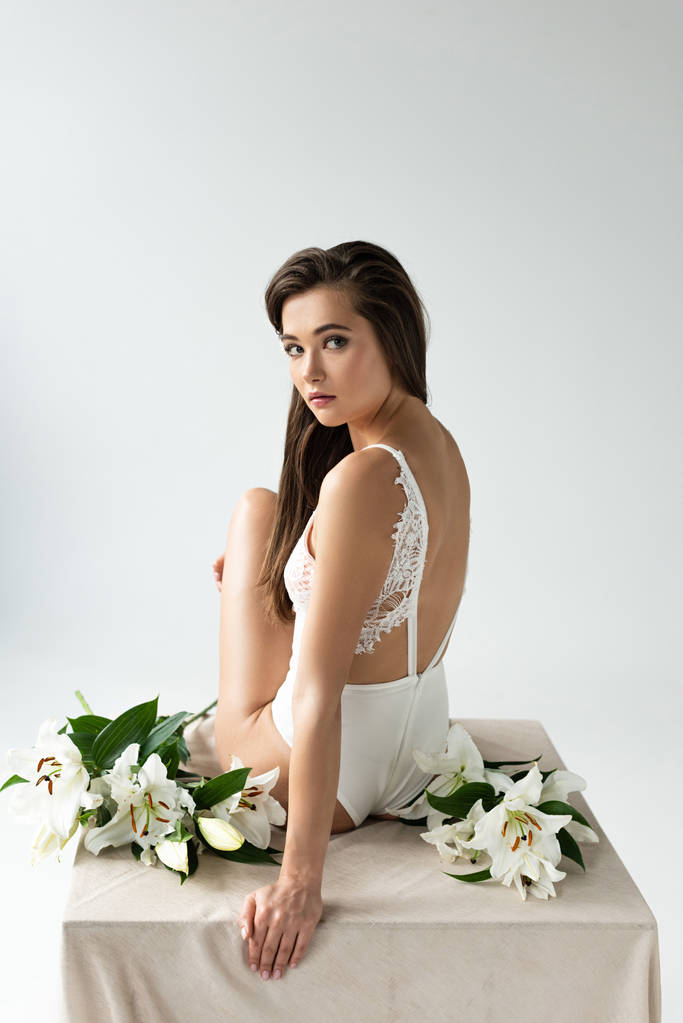 tender young woman in white lacy bodysuit sitting among lilies isolated on white - Photo, Image