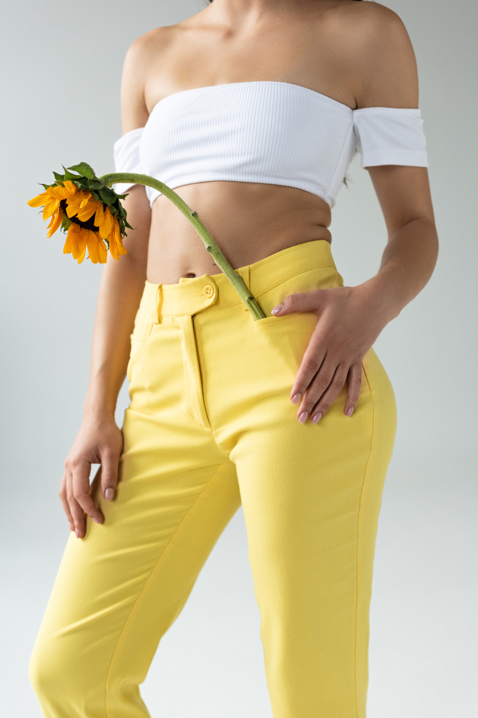 bijgesneden beeld van de vrouw in gele broek poseren met zonnebloem geïsoleerd op grijs - Foto, afbeelding