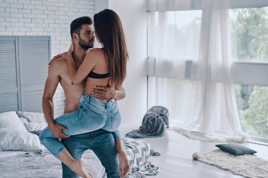 Knappe jonge shirtless man met semi-geklede aantrekkelijke vrouw terwijl ze in de slaapkamer staat - Foto, afbeelding