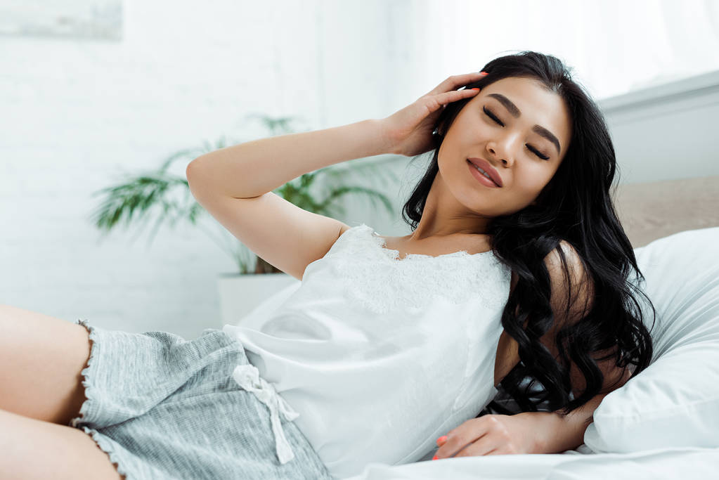 felice ragazza asiatica con gli occhi chiusi sdraiati sul letto e toccando i capelli
  - Foto, immagini