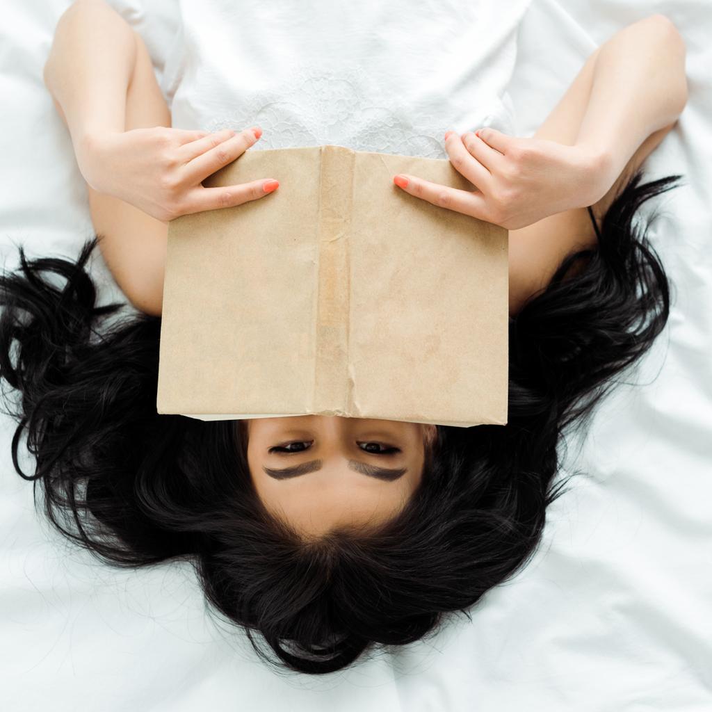 von oben: Thailänderin deckt Gesicht mit Buch ab, während sie im Bett liegt  - Foto, Bild
