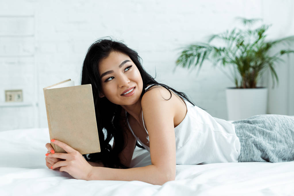 positiivinen aasialainen tyttö tilalla kirja kun makaa sängyssä kotona
  - Valokuva, kuva