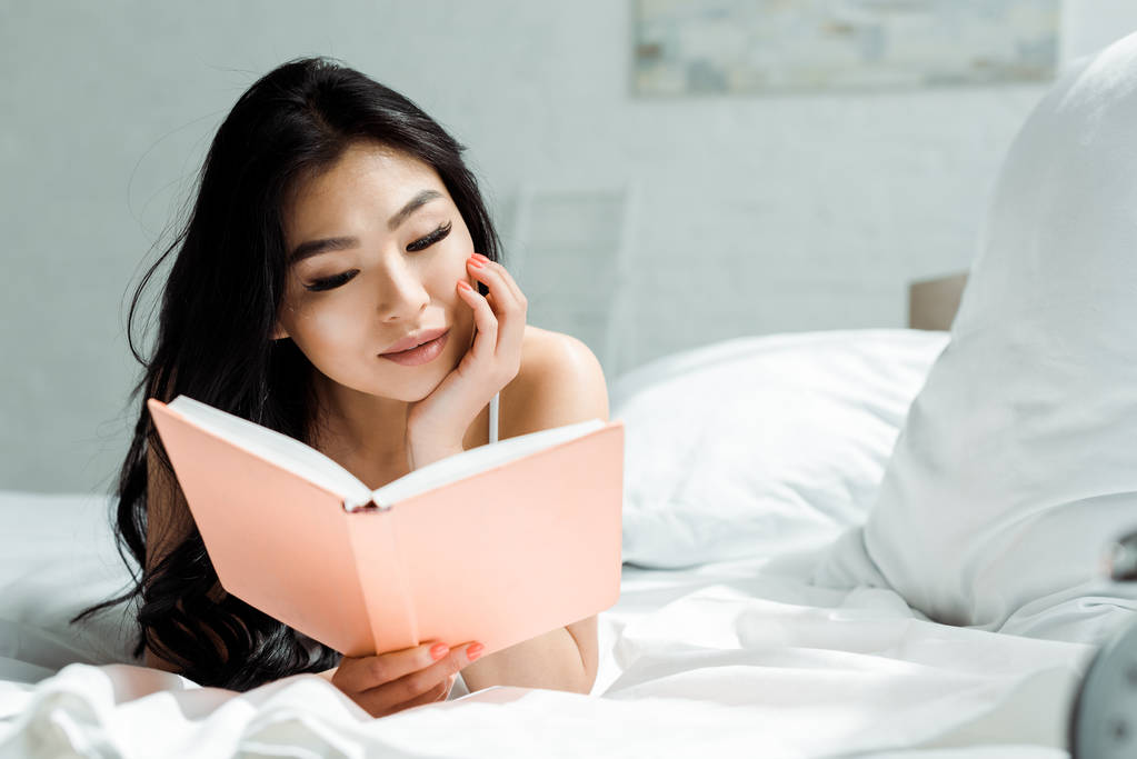 ελκυστική ταϊλανδή γυναίκα ανάγνωση βιβλίο και ξαπλωμένο στο κρεβάτι στο σπίτι  - Φωτογραφία, εικόνα