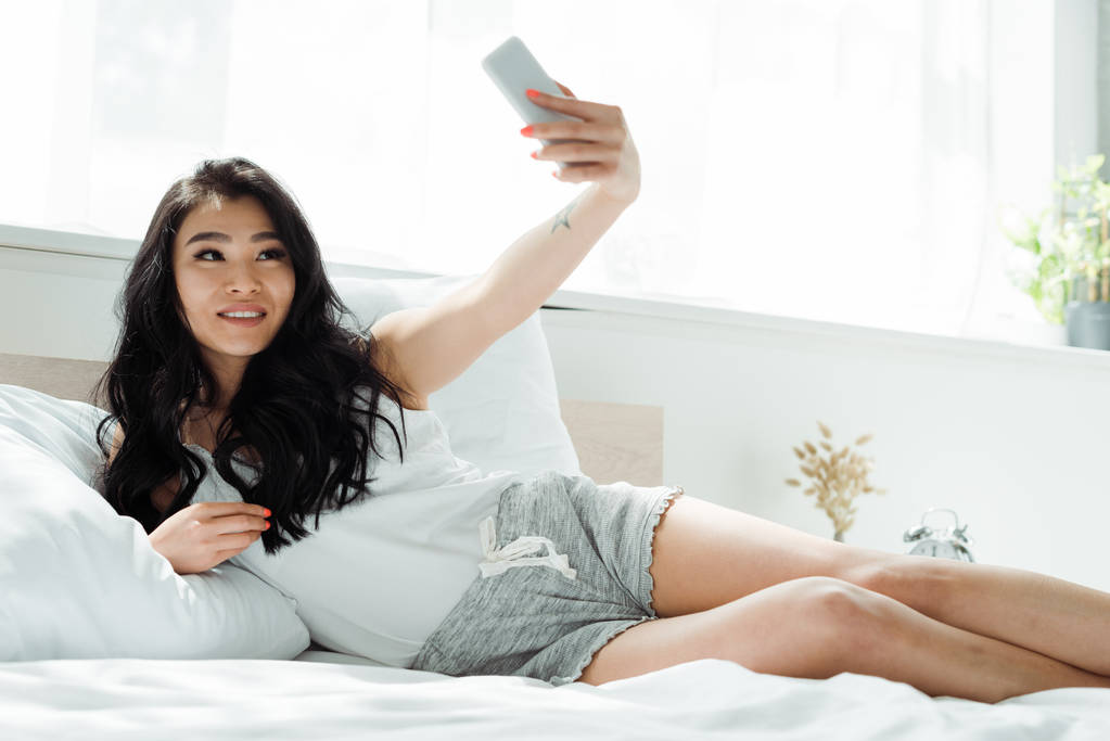 селективный фокус веселой брюнетки азиатской женщины, делающей селфи в спальне
   - Фото, изображение
