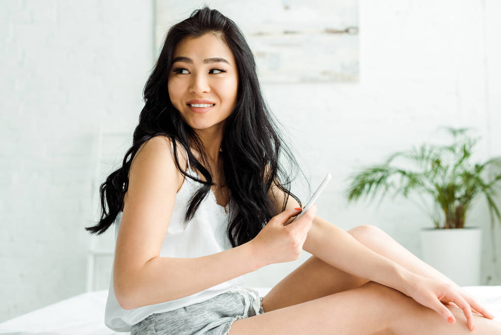 pretty thai woman holding smartphone and smiling in bedroom  - Photo, Image