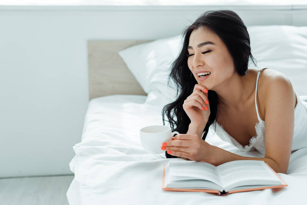 foyer sélectif de gai brunette asiatique femme couché près de livre et tenant tasse de thé
  - Photo, image