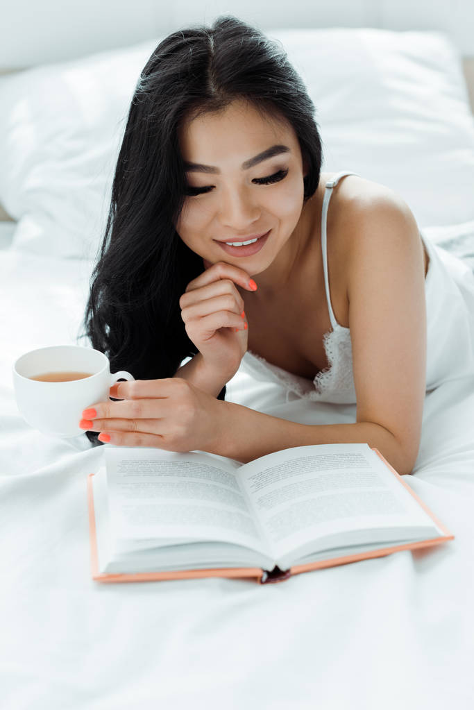 selective focus of happy brunette asian woman reading book and holding cup of tea  - Photo, Image