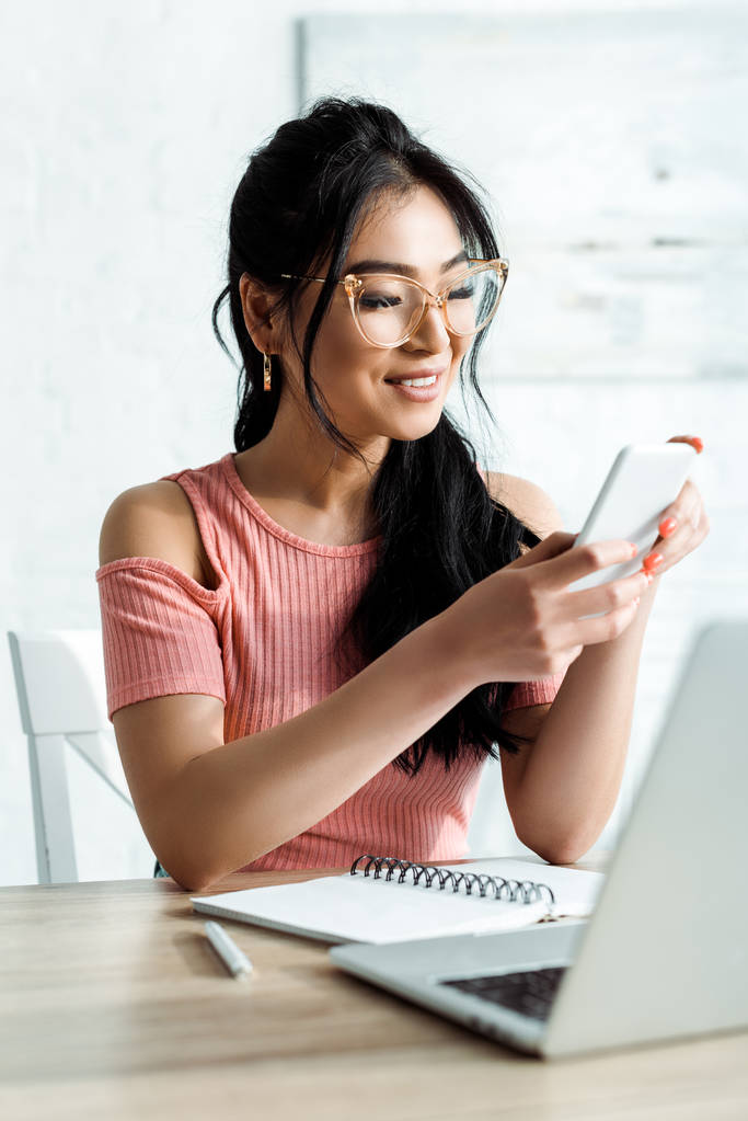 オフィスのラップトップの近くにスマートフォンを使用して眼鏡で幸せなアジアの女性の選択的な焦点  - 写真・画像