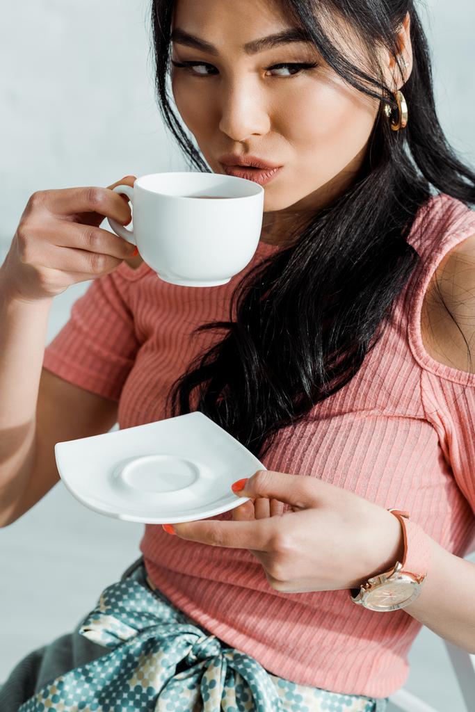 vonzó ázsiai nő gazdaság csészealj, miközben teát iszik  - Fotó, kép