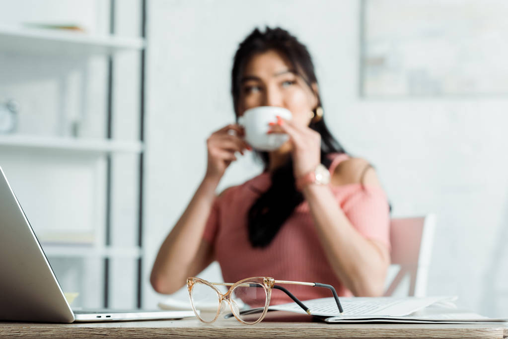 selektywnej ostrości okularów na stole w pobliżu azjatyckich kobieta picia herbaty  - Zdjęcie, obraz
