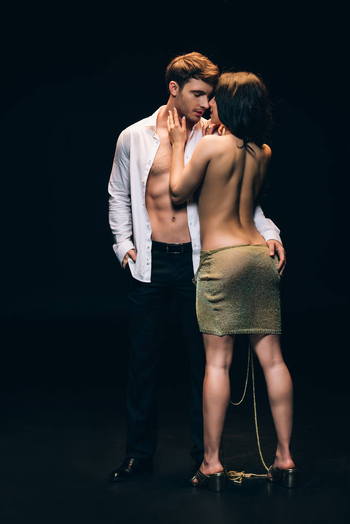 γδύσιμο γυναίκα με σέξι γλουτούς στέκεται κοντά κομψό φίλο με γυμνό κορμό απομονώνεται σε μαύρο - Φωτογραφία, εικόνα