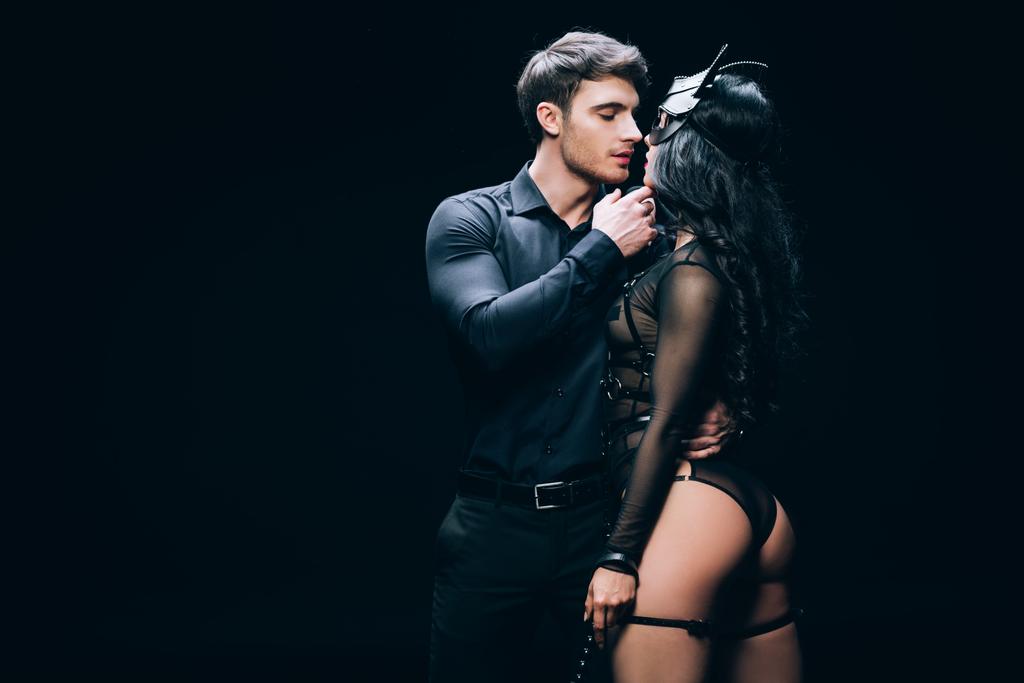 young man kissing sexy brunette woman in bdsm costume isolated on black - Photo, Image
