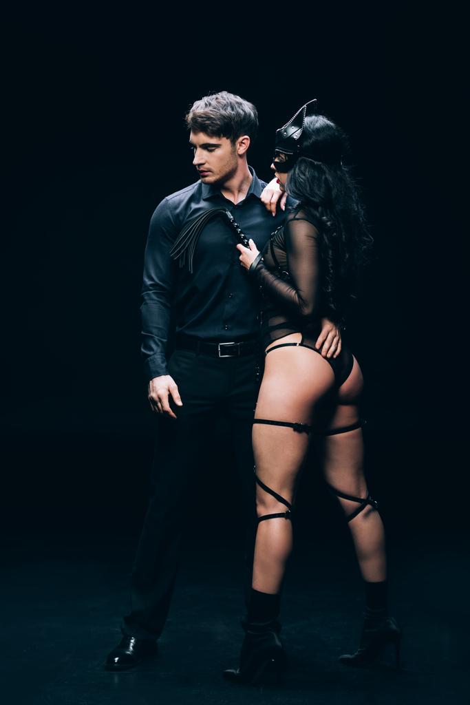 jonge man en sexy brunette vrouw in bdsm kostuum met geseling zweep op zwart - Foto, afbeelding