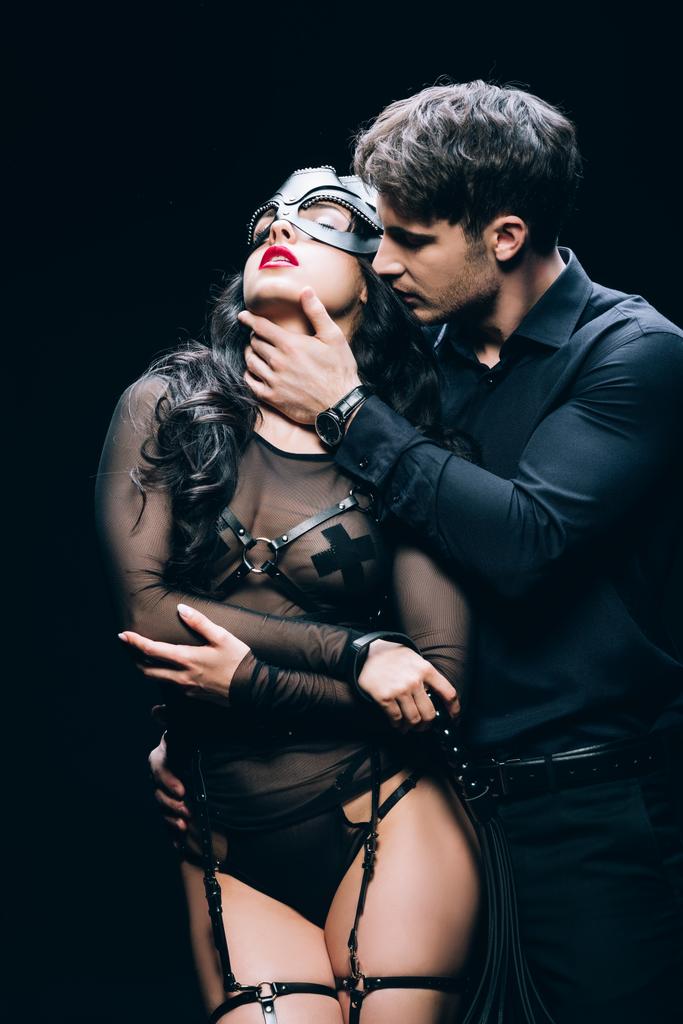 νεαρός άνδρας αγγίζει σέξι μελαχρινή γυναίκα σε bdsm κοστούμι απομονώνονται σε μαύρο - Φωτογραφία, εικόνα