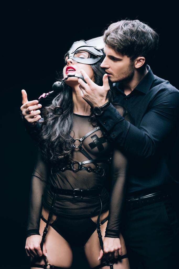 όμορφος νέος άνθρωπος κρατώντας φίμωτρο κοντά σέξι παθιασμένη γυναίκα με μάσκα και BDSM κοστούμι απομονωθεί σε μαύρο - Φωτογραφία, εικόνα