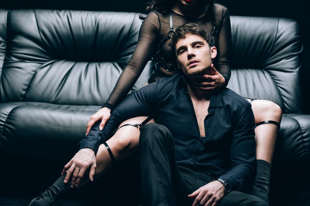 apasionado guapo hombre sentado cerca sexy mujer en bdsm traje en negro sofá de cuero
 - Foto, Imagen