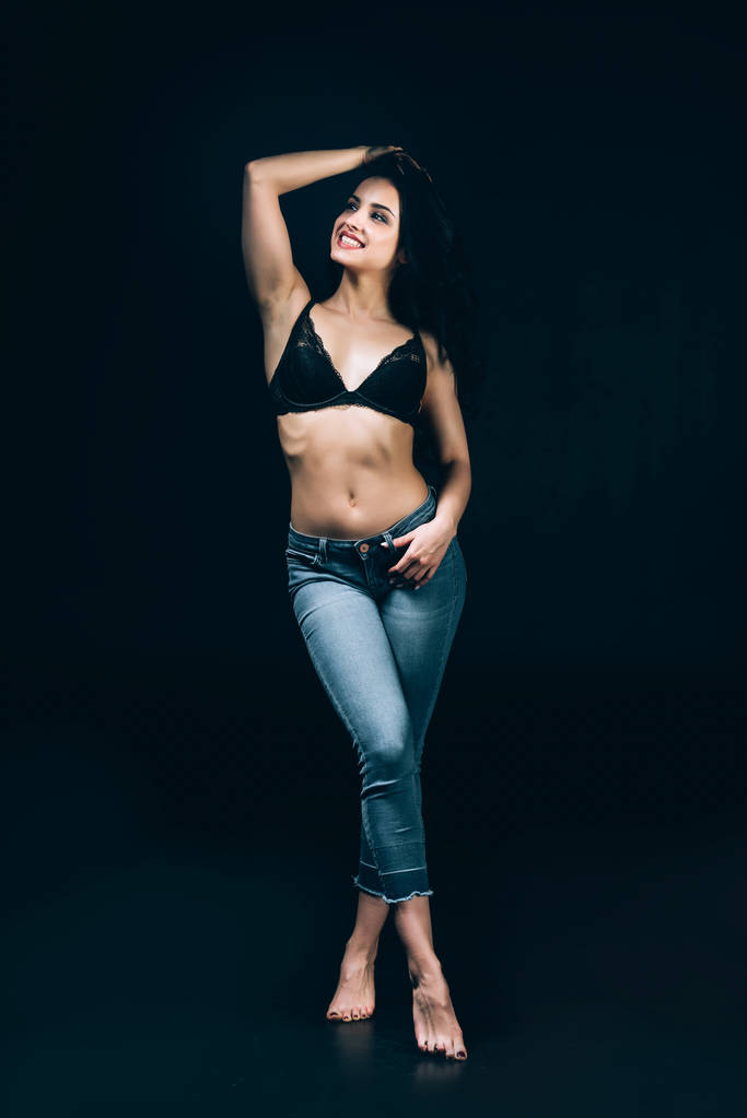 sexy feliz morena descalza mujer en azul jeans y sujetador posando aislado en negro
 - Foto, imagen