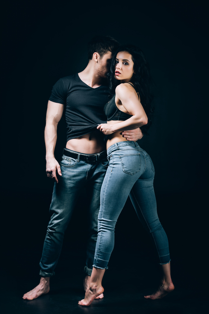 sexy barfüßiges Paar in Jeans, das sich auszieht, während es eng isoliert auf schwarz steht  - Foto, Bild