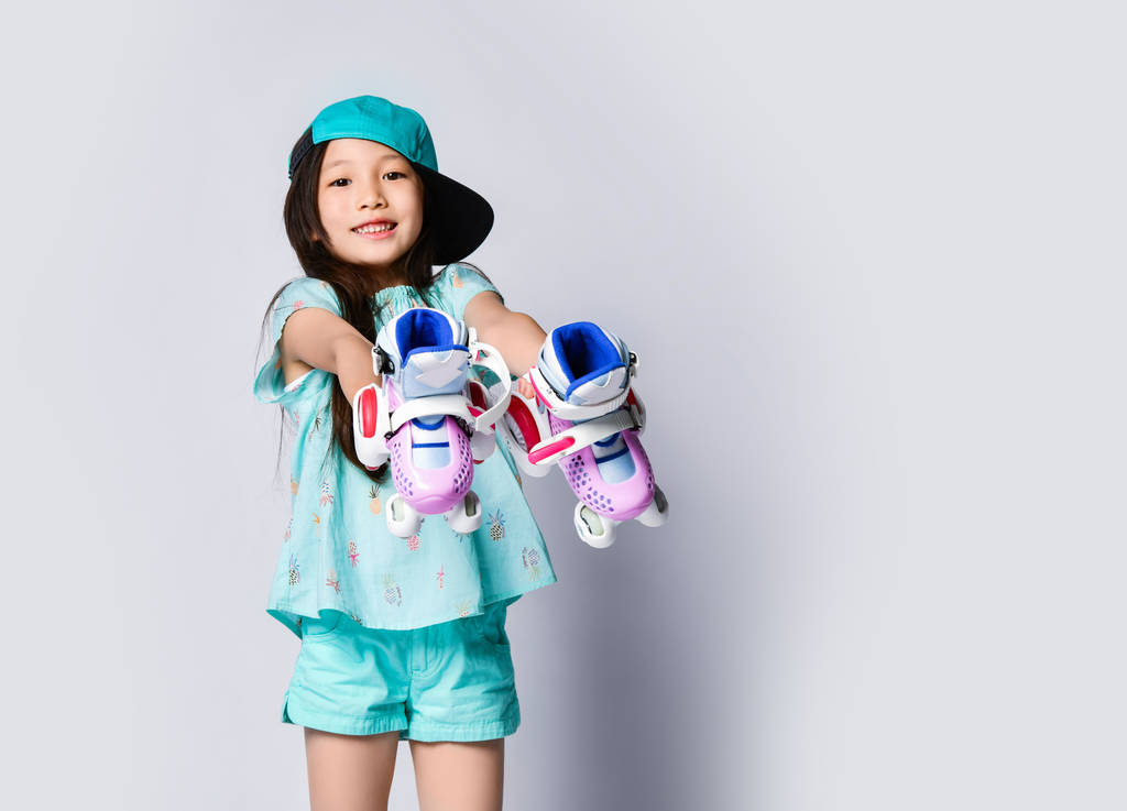 Pequeña niña asiática sentada con patines en camiseta azul claro y gorra feliz sonriendo en blanco
 - Foto, imagen