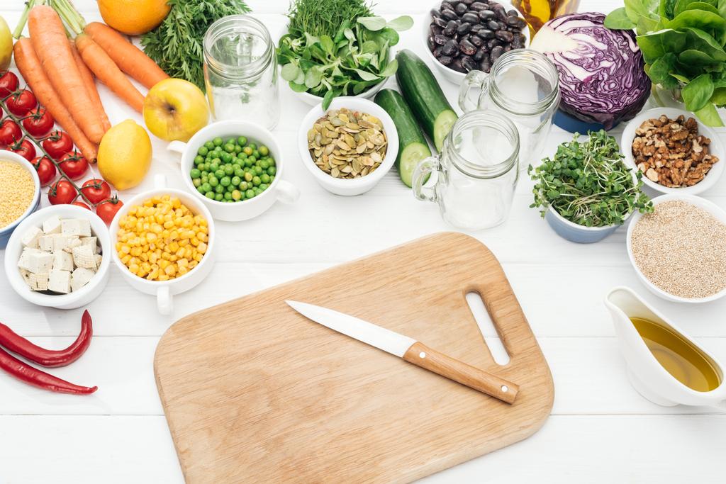 vista superior de frutas y verduras frescas cerca de frascos de vidrio en la mesa blanca de madera con tabla de cortar y cuchillo
 - Foto, Imagen