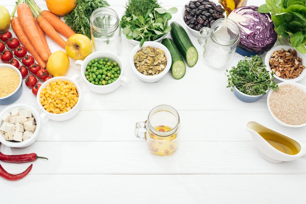 pohled na skleněnou nádobu s olejem a chilli paprik v blízkosti plodů a zeleniny v blízkosti skleniček na dřevěném bílém stole - Fotografie, Obrázek