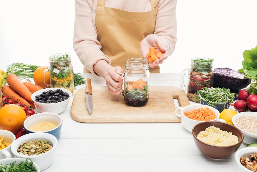 osittainen näkymä nainen esiliina lisäämällä viipaloitu porkkana purkissa salaatti puinen pöytä eristetty valkoinen
 - Valokuva, kuva