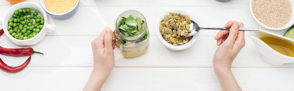 vue recadrée de femme ajoutant des graines de citrouille dans un bocal avec salade sur une table blanche en bois, vue panoramique
 - Photo, image