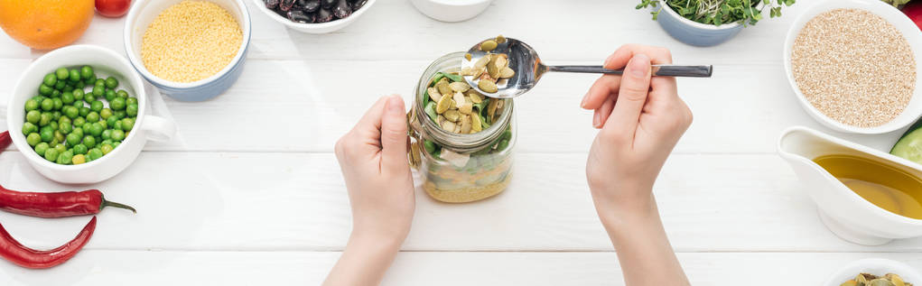 partial view of woman adding pumpkin seeds in jar with salad on wooden white table, panoramic shot - Photo, Image