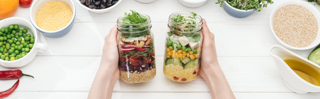 cropped view of woman holding jars with tasty salad on wooden white table , panoramic shot - Photo, Image