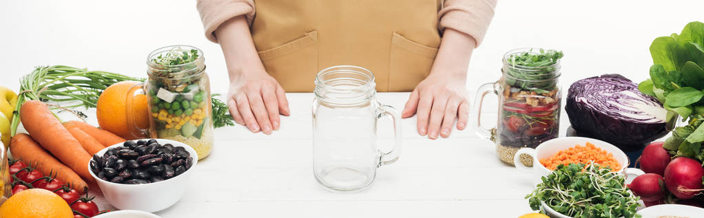 vista recortada de la mujer en el delantal de pie cerca de frasco de vidrio vacío en la mesa de madera aislado en blanco, tiro panorámico
 - Foto, Imagen