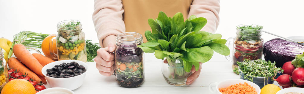 geschnittene Ansicht einer Frau in Schürze, die ein Glas mit Salat und grünen Blättern auf einem Holztisch hält, isoliert auf weißem Hintergrund, Panoramaaufnahme - Foto, Bild