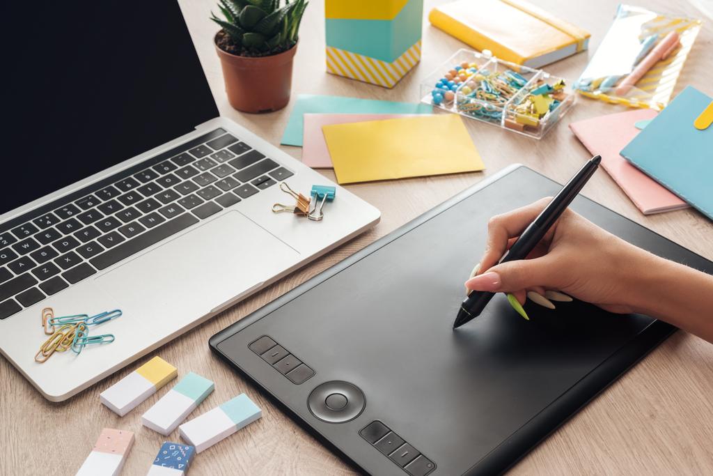 oříznutý pohled na ženu pracující s kreslicím tabletem, sedící za dřevěnou tabulkou s papírnickým stolem a laptopem - Fotografie, Obrázek