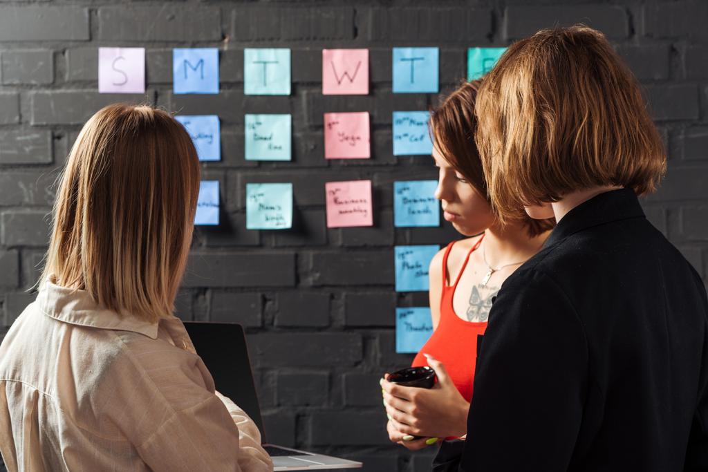 women standing near colorful sticker notes on black brick wall, looking at laptop - Photo, Image