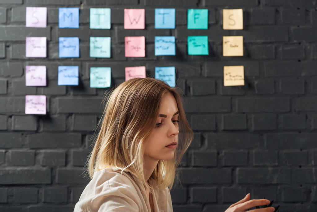 side view of woman standing near colorful sticker notes on black brick wall, looking away - Photo, Image