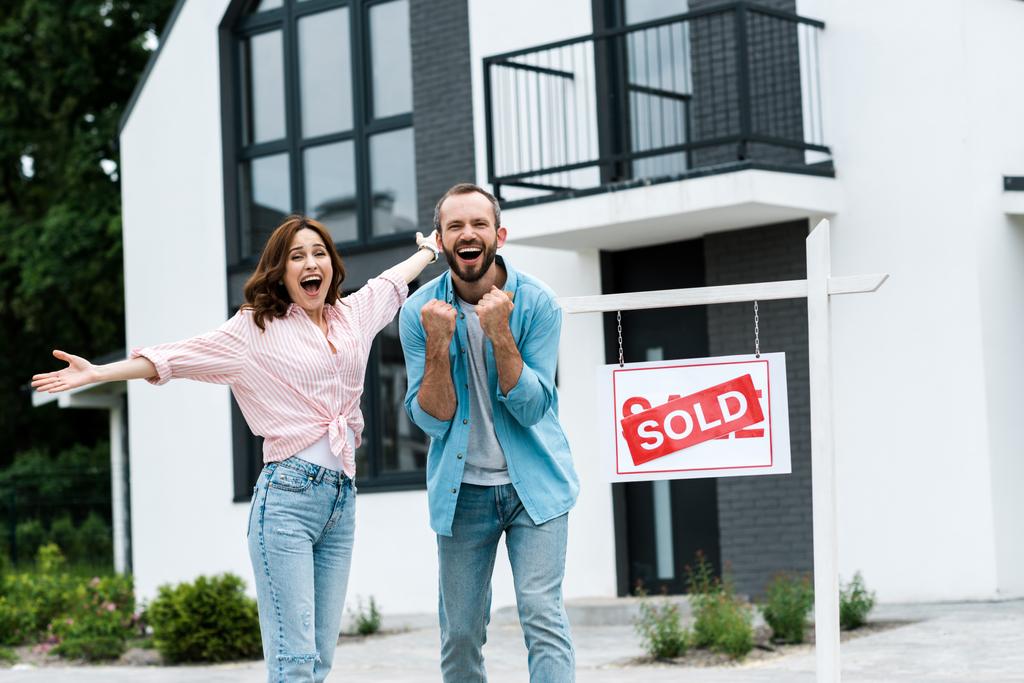 cheerful man and woman celebrating near house and board with sold letters  - Photo, Image