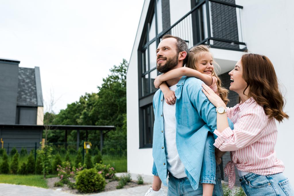 happy man piggybacking daughter near cheerful wife and house  - Photo, Image